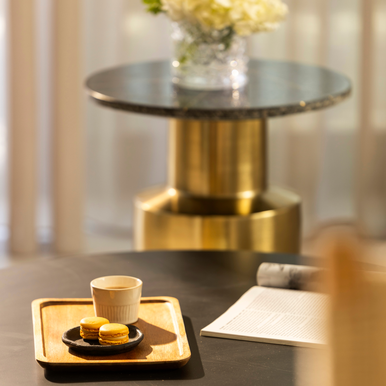 Cozy cafe scene: macarons and coffee on a wooden tray, open magazine, brass pedestal table with flowers in soft focus.