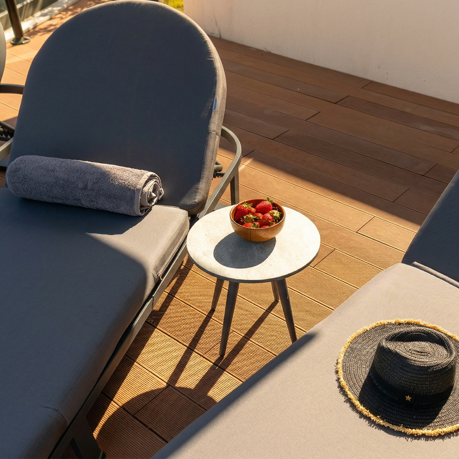 Sunlit outdoor terrace with gray lounge chairs, a rolled towel, and a small table of fresh strawberries.