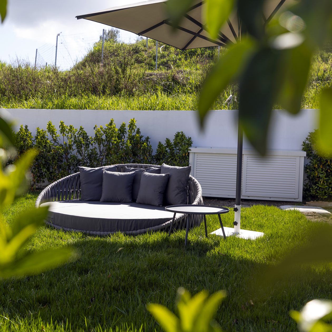 Outdoor round wicker daybed with gray cushions and umbrella on a green lawn with hedge backdrop.