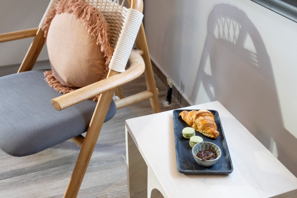Cozy minimalist cafe scene: croissant, two green macarons and jam on a slate plate beside a wooden chair.