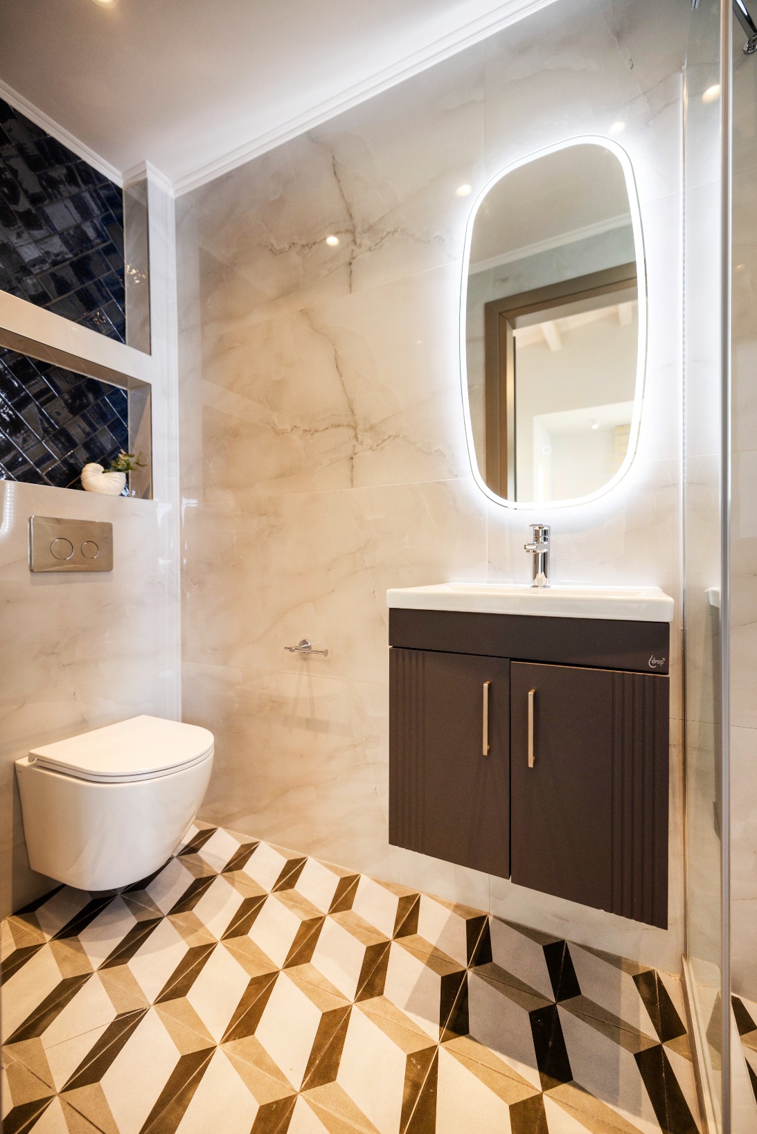 Modern bathroom with wall-mounted toilet, floating dark vanity, backlit oval mirror, marble walls, and geometric floor tiles.
