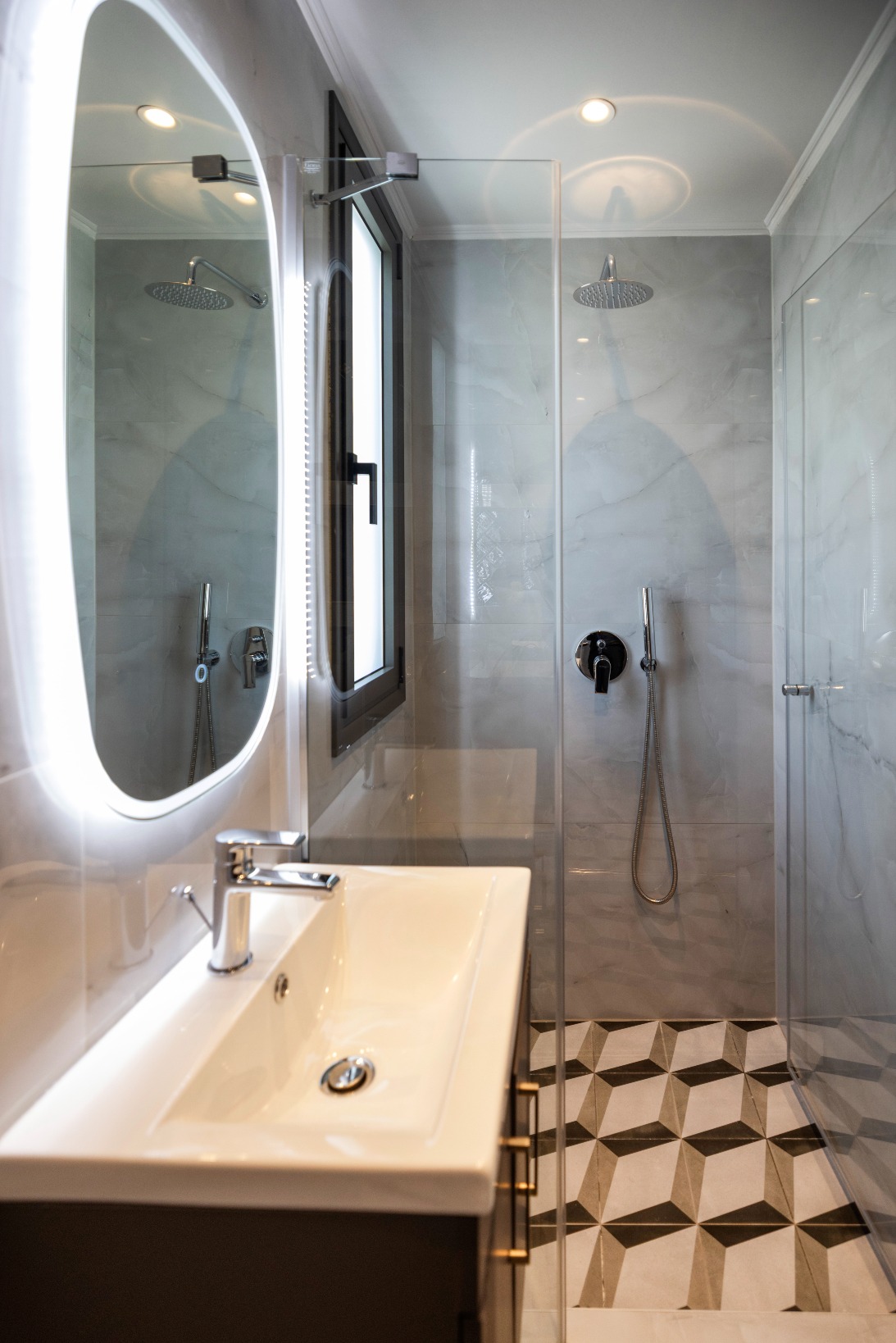 Modern bathroom with glass shower enclosure, round LED mirror, white vanity sink, marble walls, and geometric floor tiles.
