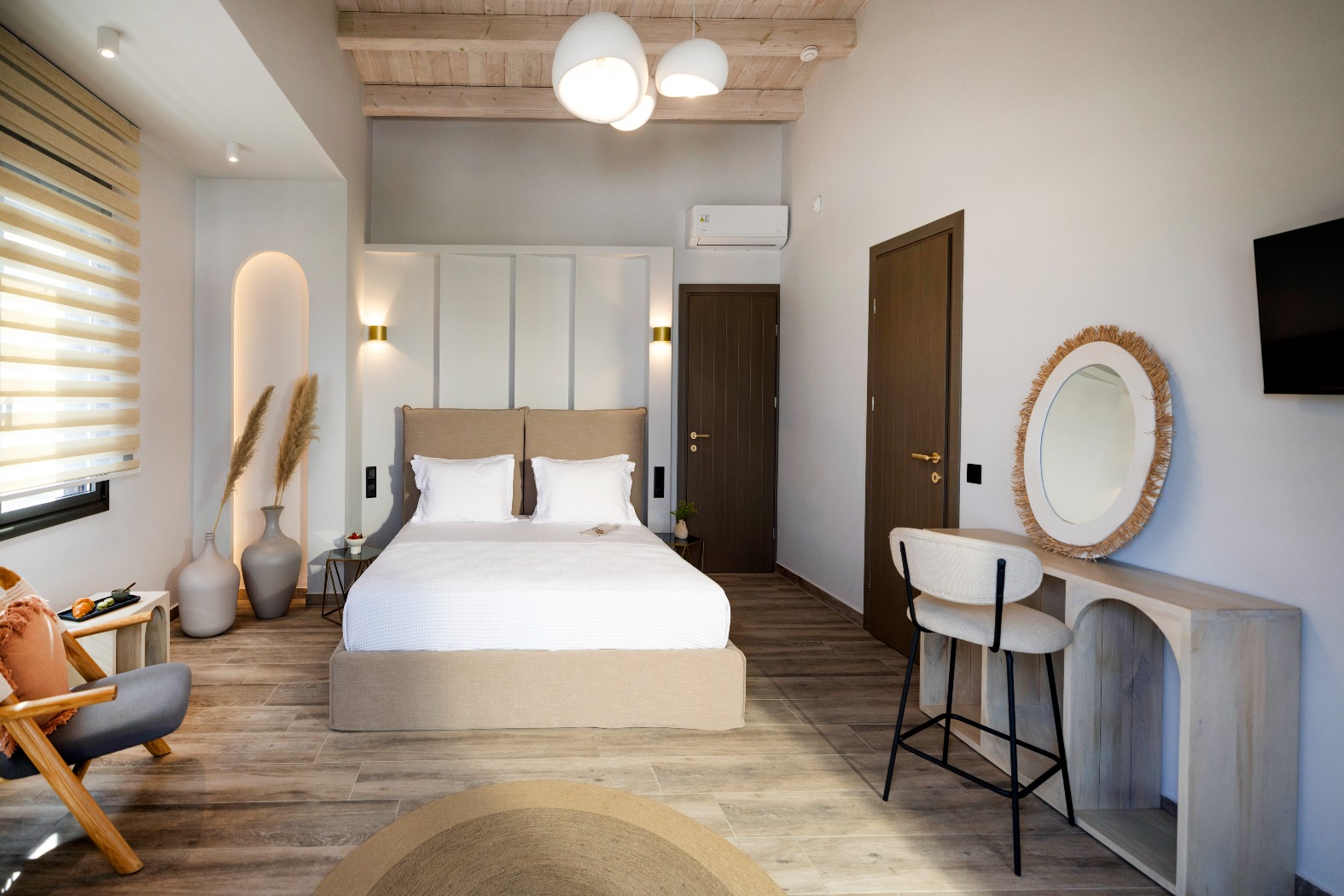 Modern hotel room with king bed, beige headboard, wooden ceiling beams, pampas decor, TV and straw-framed mirror.