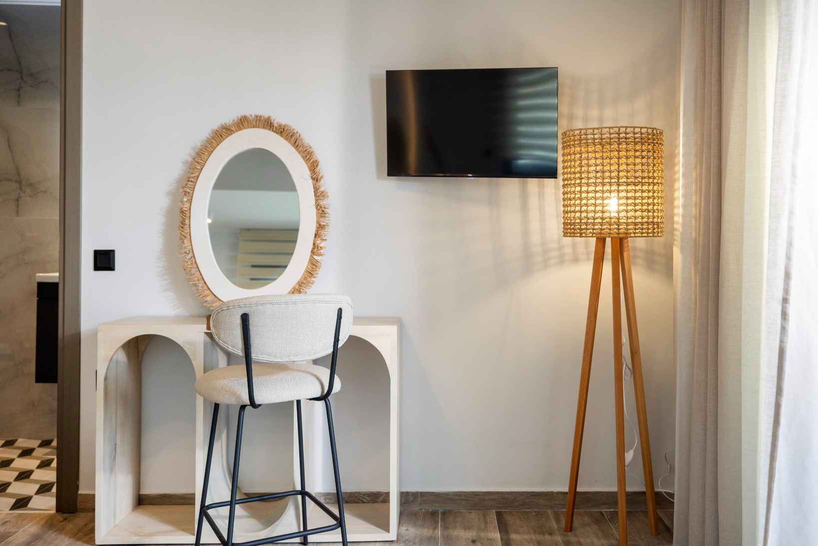 Modern dressing area: round fringed straw mirror, white vanity, beige stool, wall-mounted TV, woven tripod lamp.