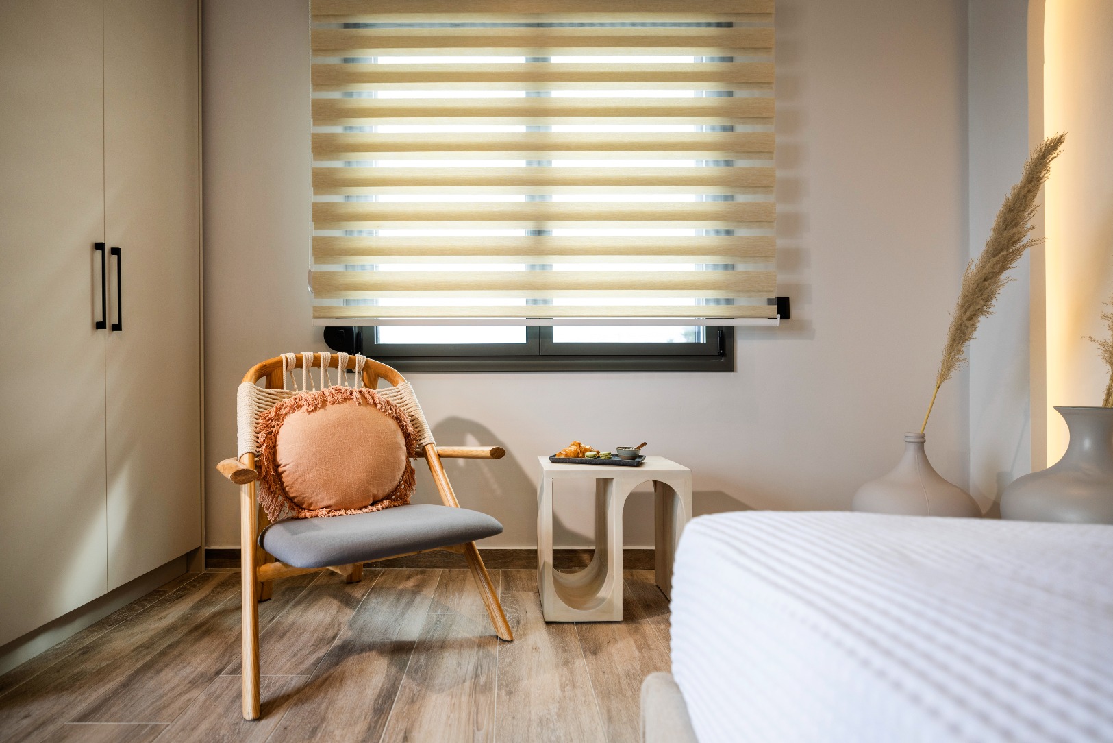 Cozy modern bedroom corner with wicker chair, beige striped blinds, small side table with croissants, pampas grass vase.
