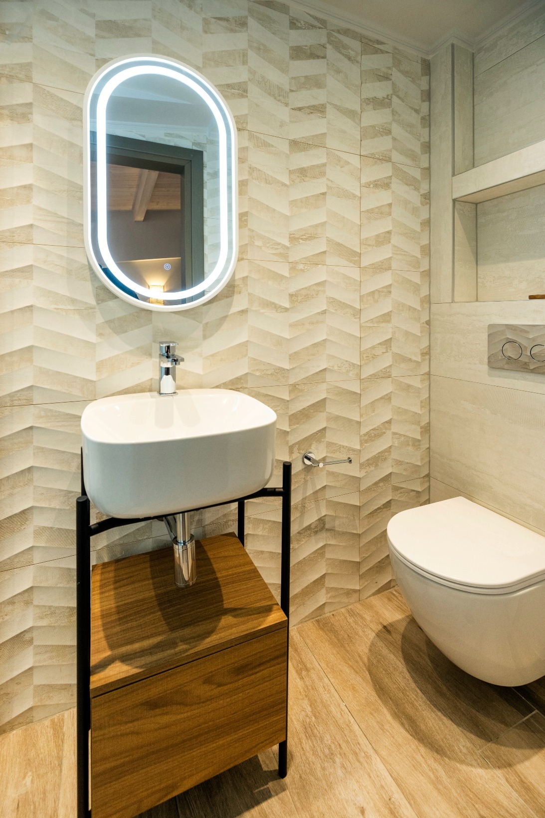 Modern bathroom with LED-ring oval mirror, white vessel sink on a wooden cabinet with black frame, and wall-mounted toilet.