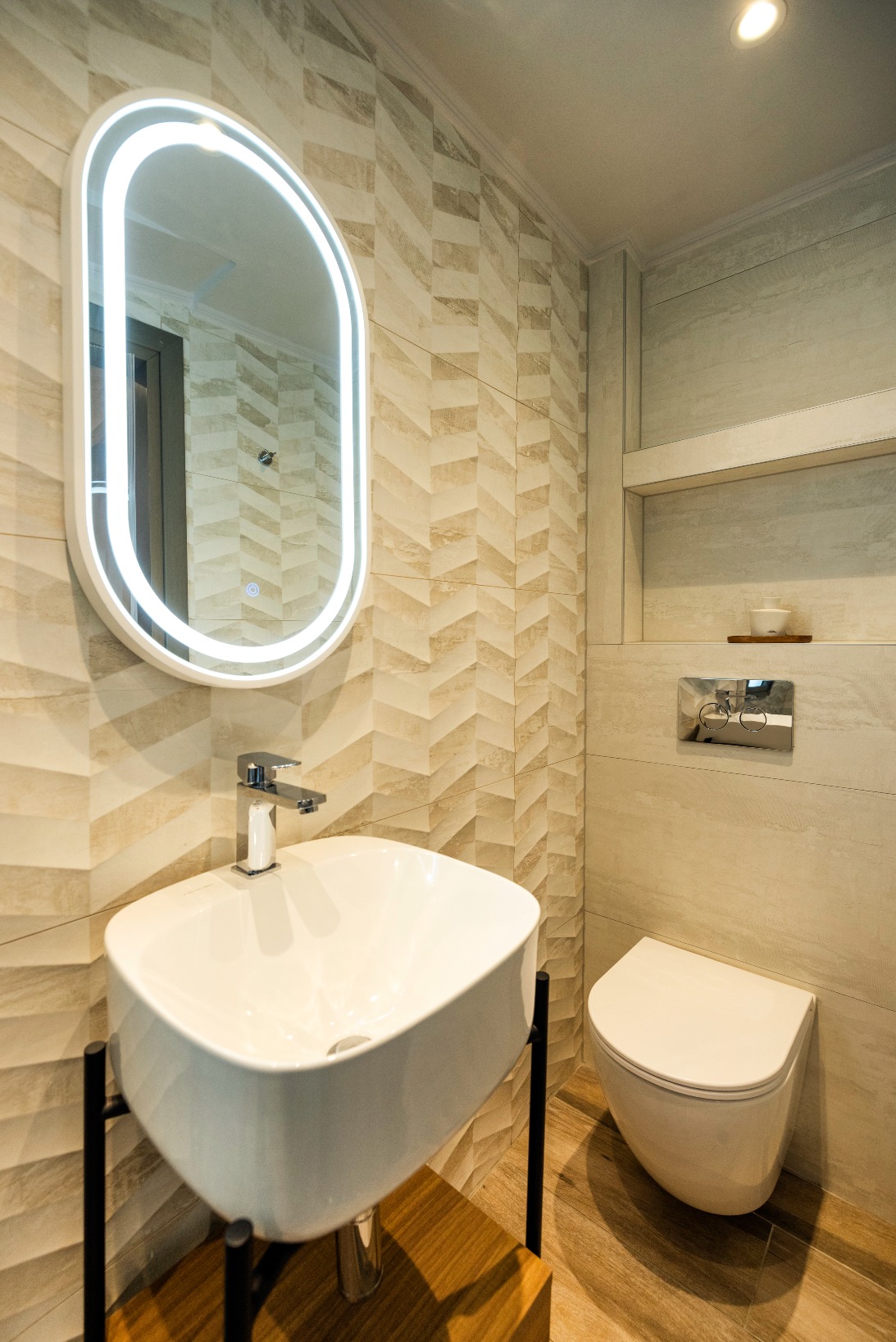 Modern bathroom with LED-illuminated oval mirror, white sink on black stand, and wall-hung toilet.