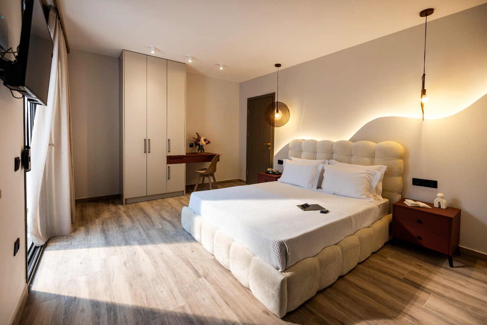 Modern hotel room with king bed, tufted beige headboard, wardrobe, desk, warm pendant lights, and wooden floor.