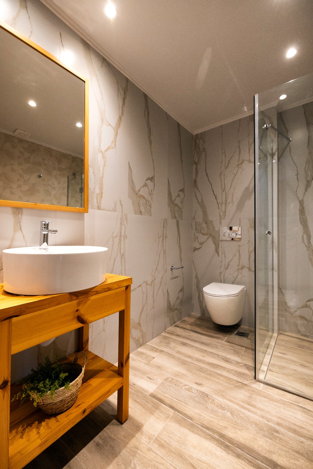 Modern bathroom with marble walls, wooden vanity, round vessel sink, gold-framed mirror, glass shower and wall-hung toilet.