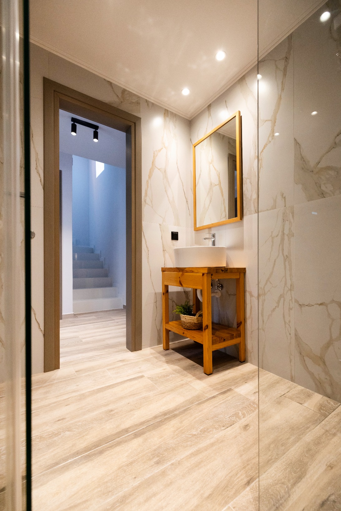 Modern bathroom with marble walls, wooden vanity, vessel sink and gold-framed mirror.