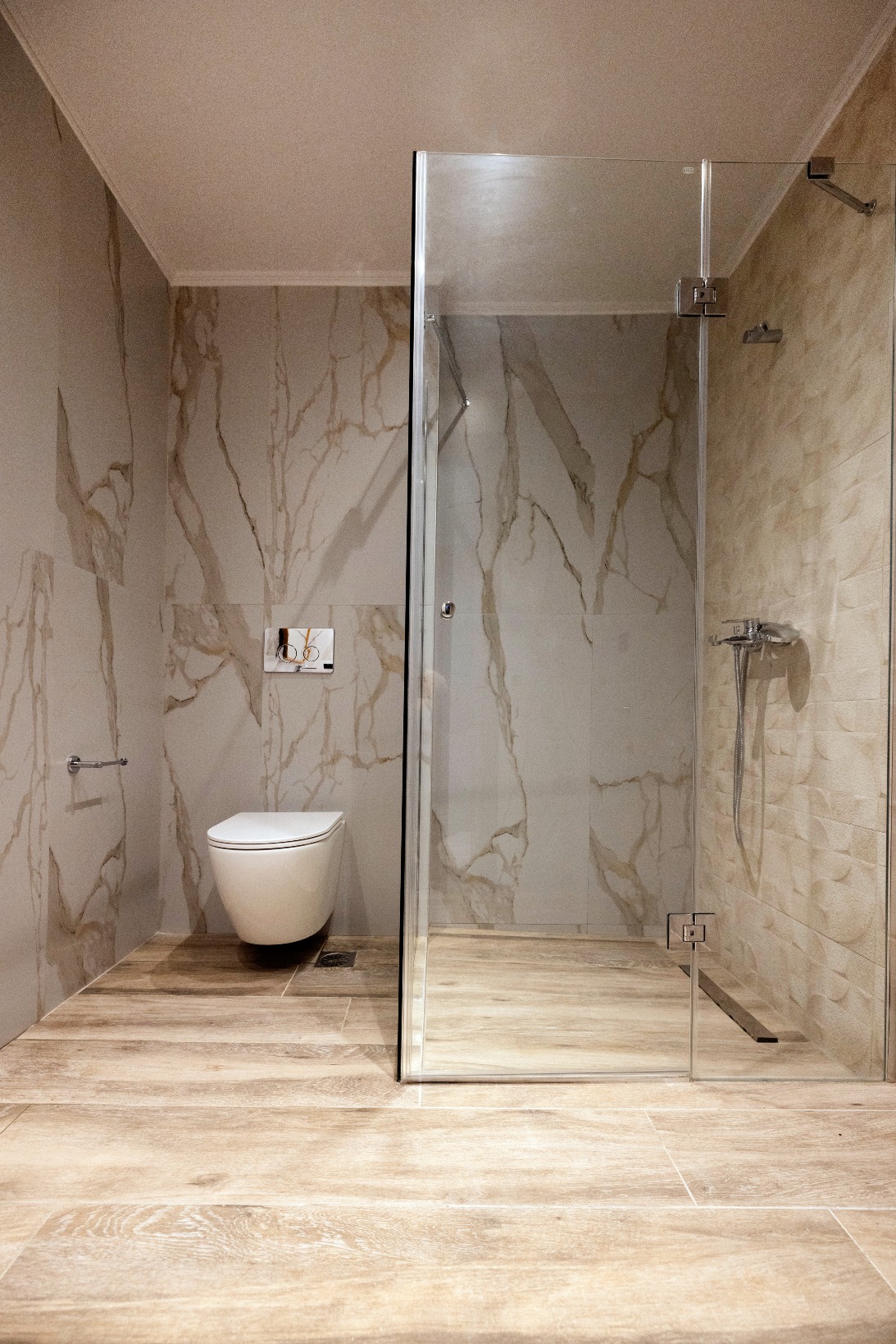 Modern marble bathroom with glass shower enclosure, wall-mounted toilet, and wood-look beige floor tiles.