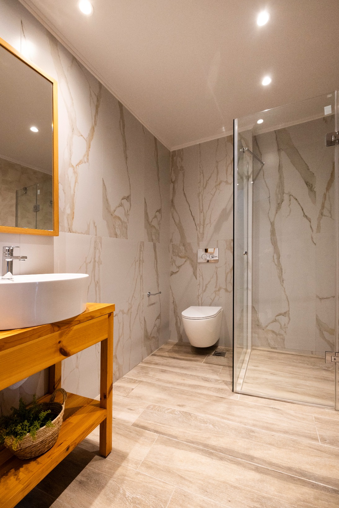 Bright modern bathroom with marble-tile walls, glass shower, wall-mounted toilet, and wooden vanity with vessel sink.