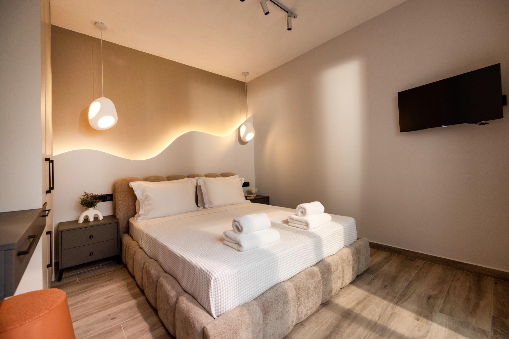 Modern hotel bedroom with a king bed, wave-lit headboard, pendant lights, wall-mounted TV, and neatly folded towels.
