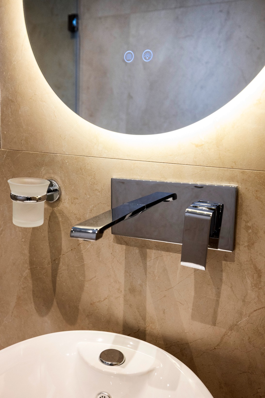 Round backlit bathroom mirror over a sleek chrome wall-mounted faucet and white sink.