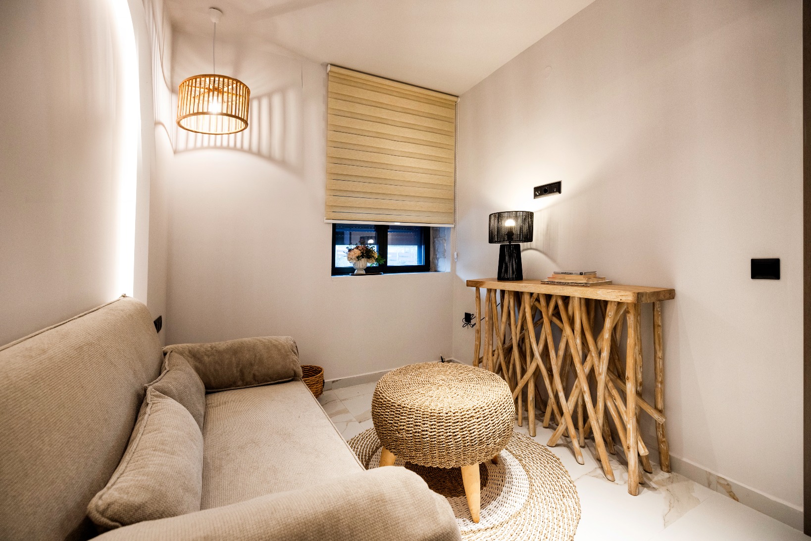 Cozy living nook with beige sofa, woven pouf, natural wood console, warm pendant light, and window with roman shade.