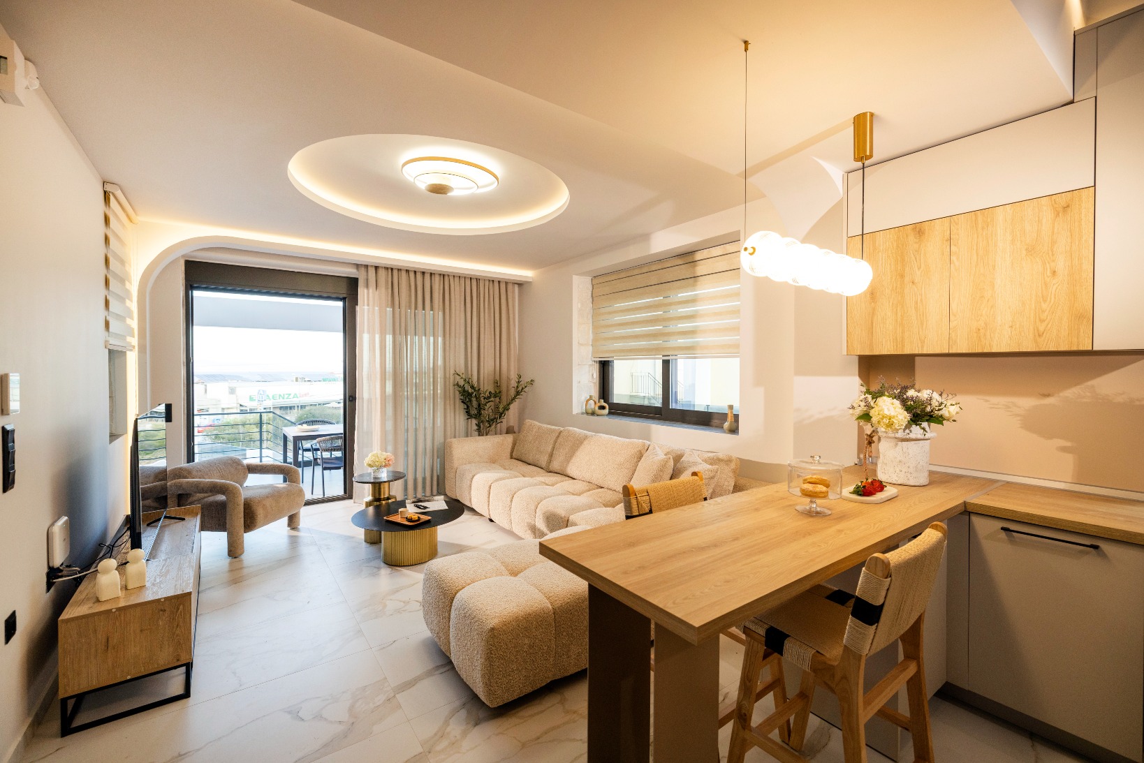 Modern living room with beige sectional, open-plan kitchen, marble floor, balcony view and chic pendant lighting.