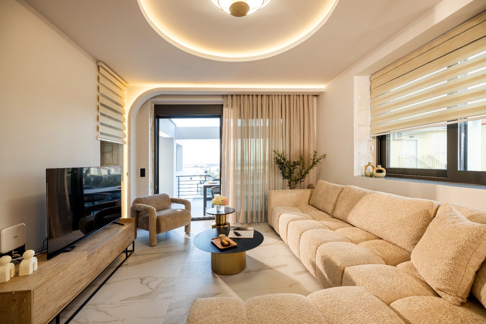 Bright modern living room with beige sectional sofa, marble floor, TV console, and balcony with sheer curtains.