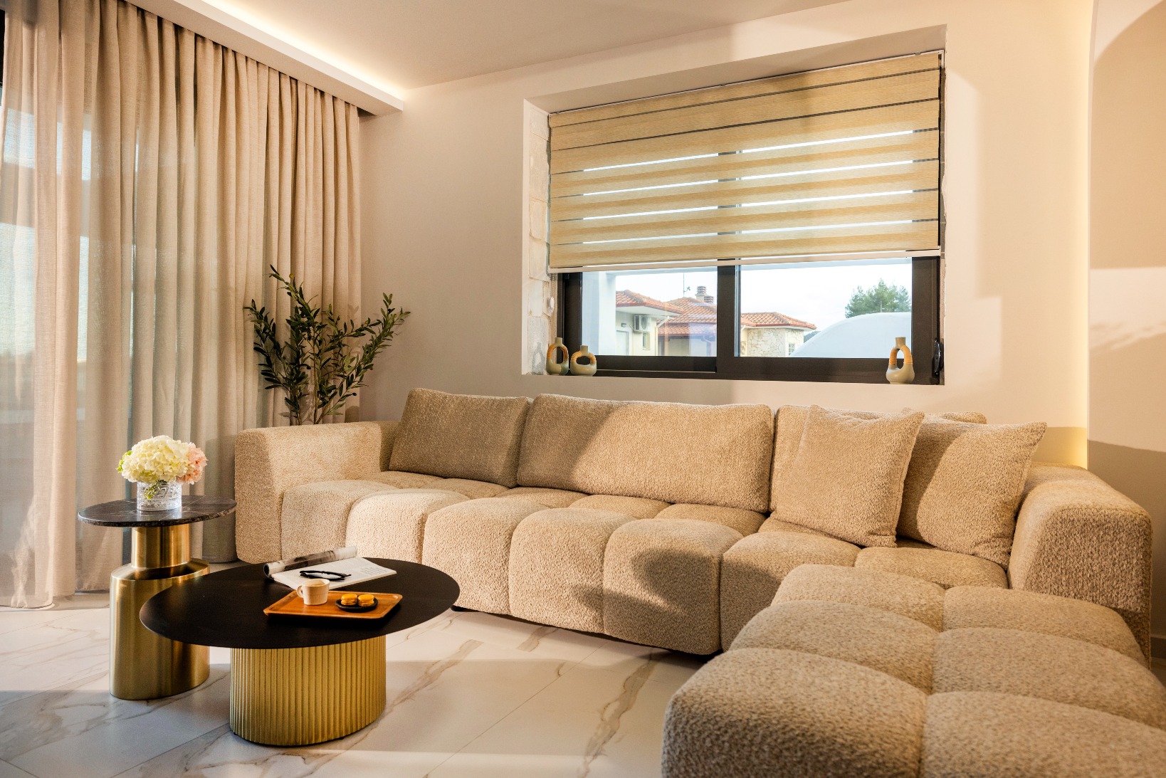 Modern beige sectional sofa in a bright living room with gold coffee tables and sheer curtains.