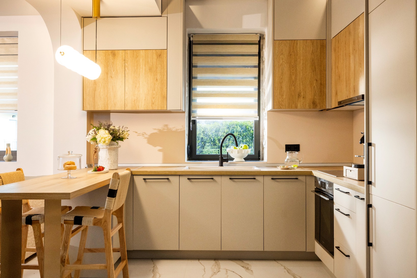 Modern kitchen with light wood cabinets, beige countertops, central island/dining table, and a window with striped roller blind.
