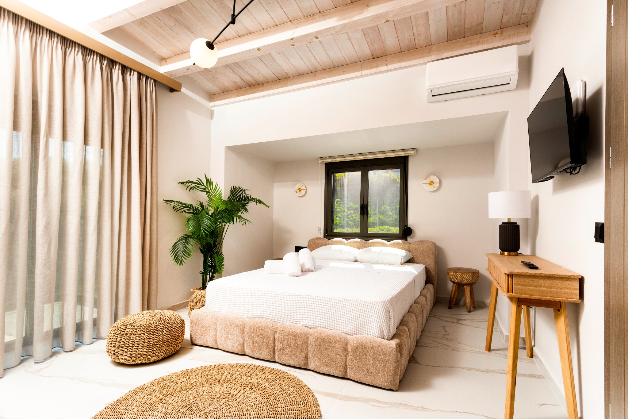Bright hotel bedroom with vaulted wooden ceiling, beige curtains, king bed, desk, plant, and wall-mounted TV.