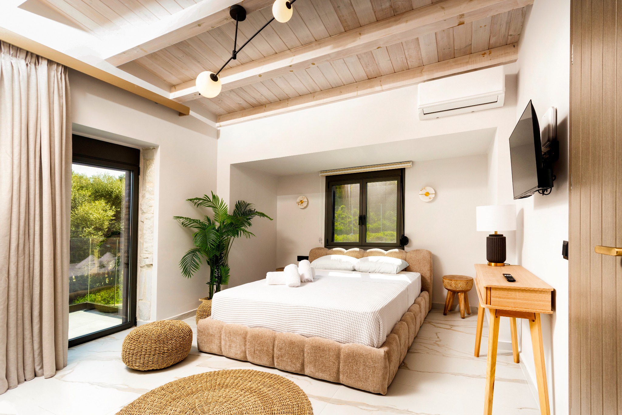 Bright modern hotel bedroom with exposed wooden beam ceiling, large bed, plant, and wall-mounted TV.