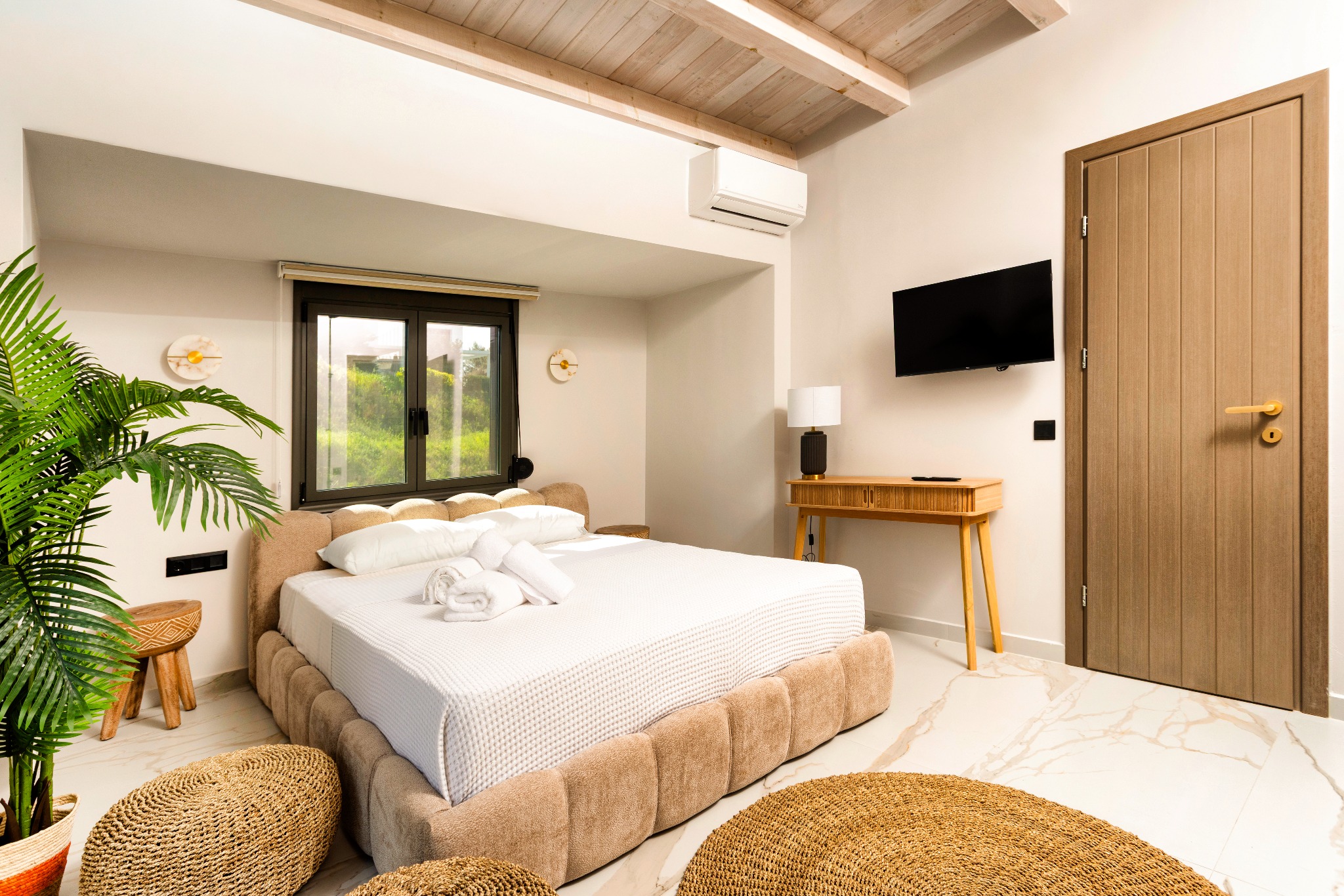 Modern bedroom with beige tufted bed, wicker poufs, potted plant, wall-mounted TV, window, and wooden door.