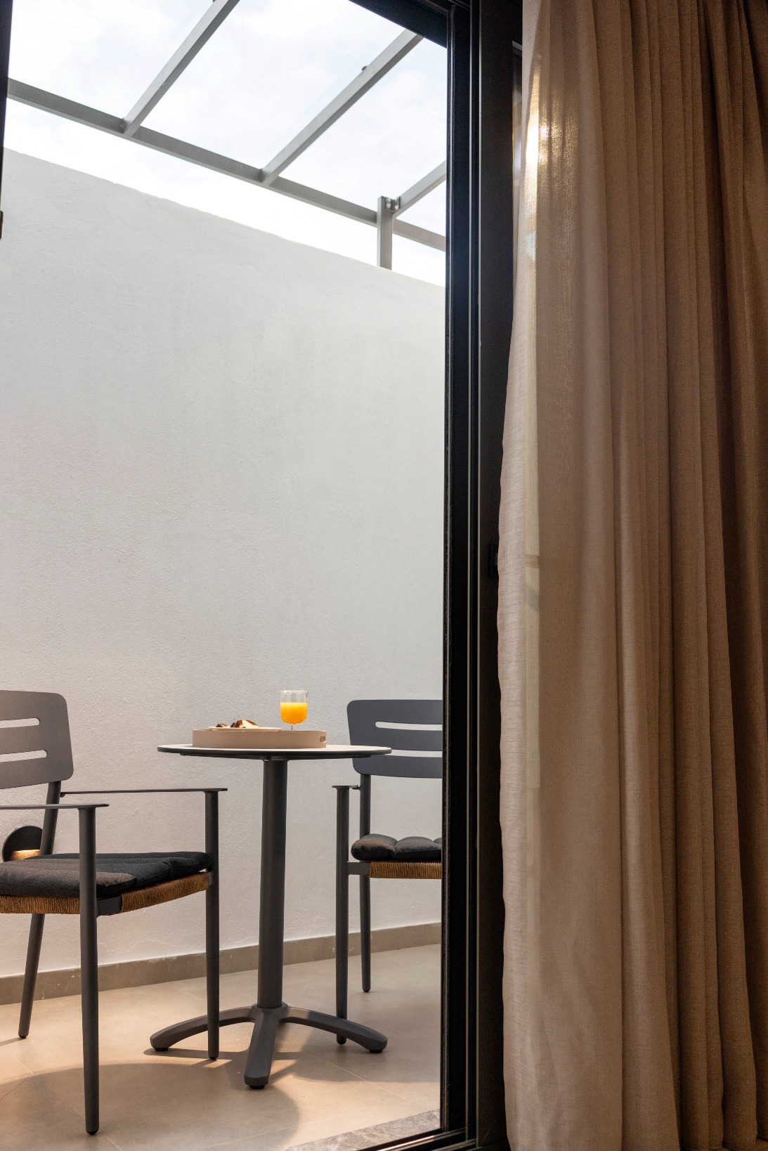 Sunlit outdoor patio table with pastries and orange juice, two chairs, beige-curtained glass door.