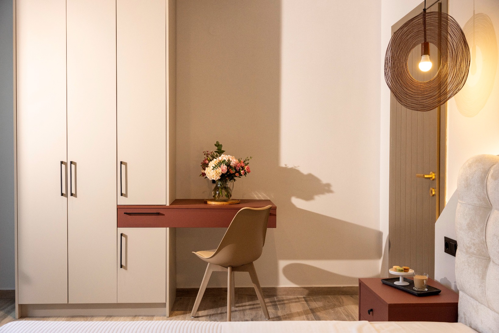 Modern bedroom workspace with white wardrobe, pink desk, vase of flowers, beige chair, and woven pendant light.