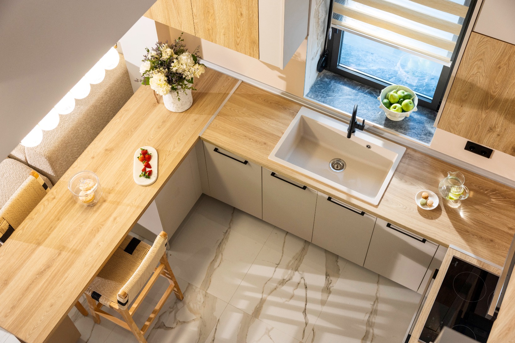 Modern kitchen seen from above with light wood worktops, white sink, a vase of flowers, plates of fruit, a glass of water, and a bowl of apples on the windowsill. Marble floor and a wooden chair are visible.