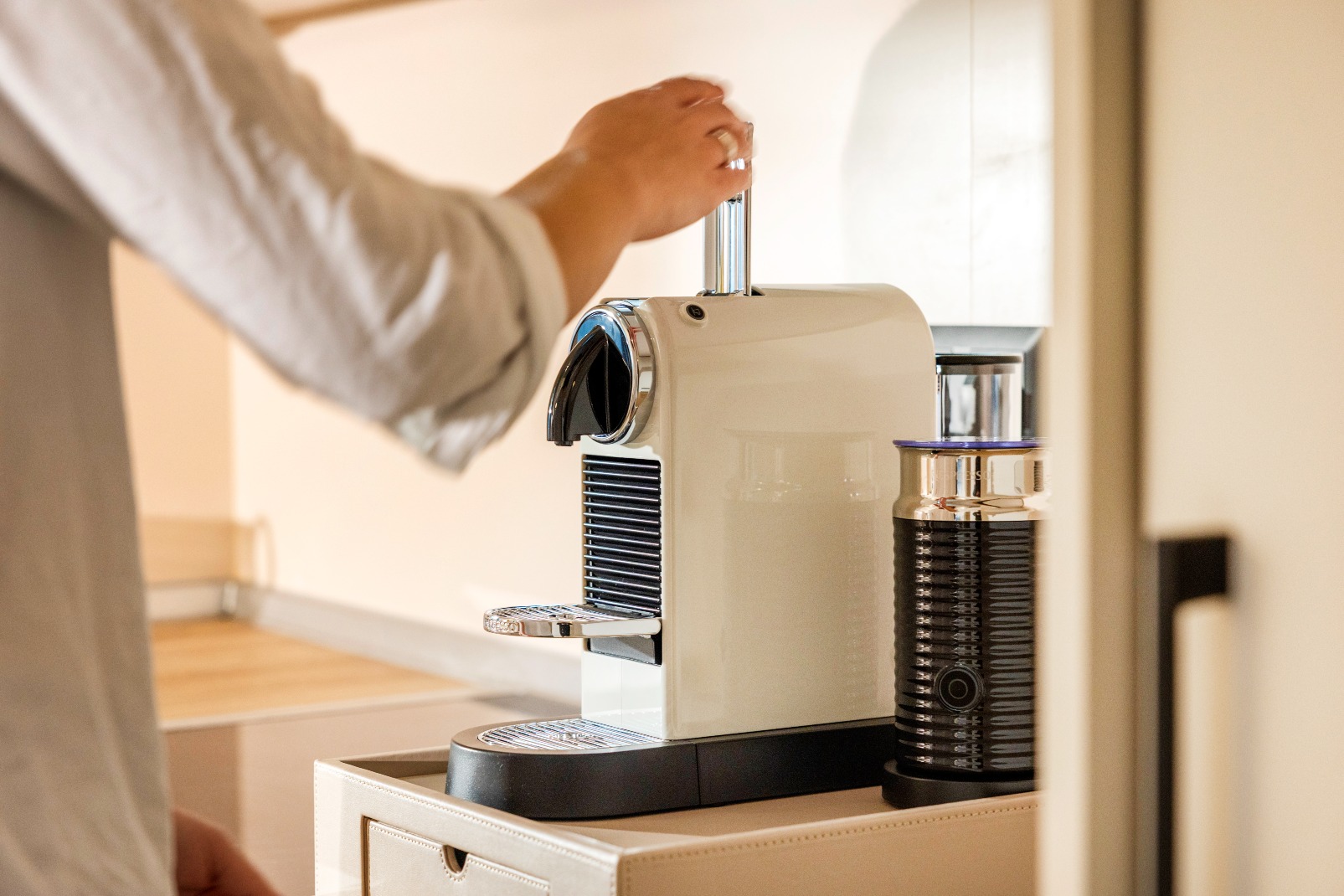 A person’s hand operates a modern white espresso machine on a kitchen worktop, with a milk frother placed beside it. The scene suggests coffee preparation in a home environment.