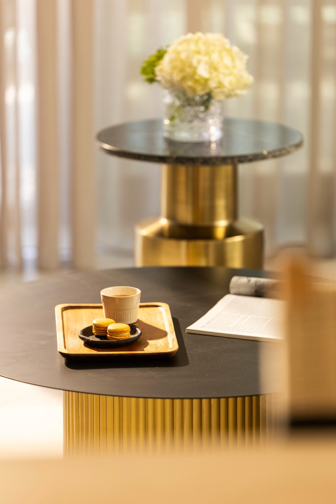 A black coffee table with a gold base holds a wooden tray with a cup, saucer, and macarons. In the background, there is a smaller table with a vase of white flowers in front of sheer curtains.