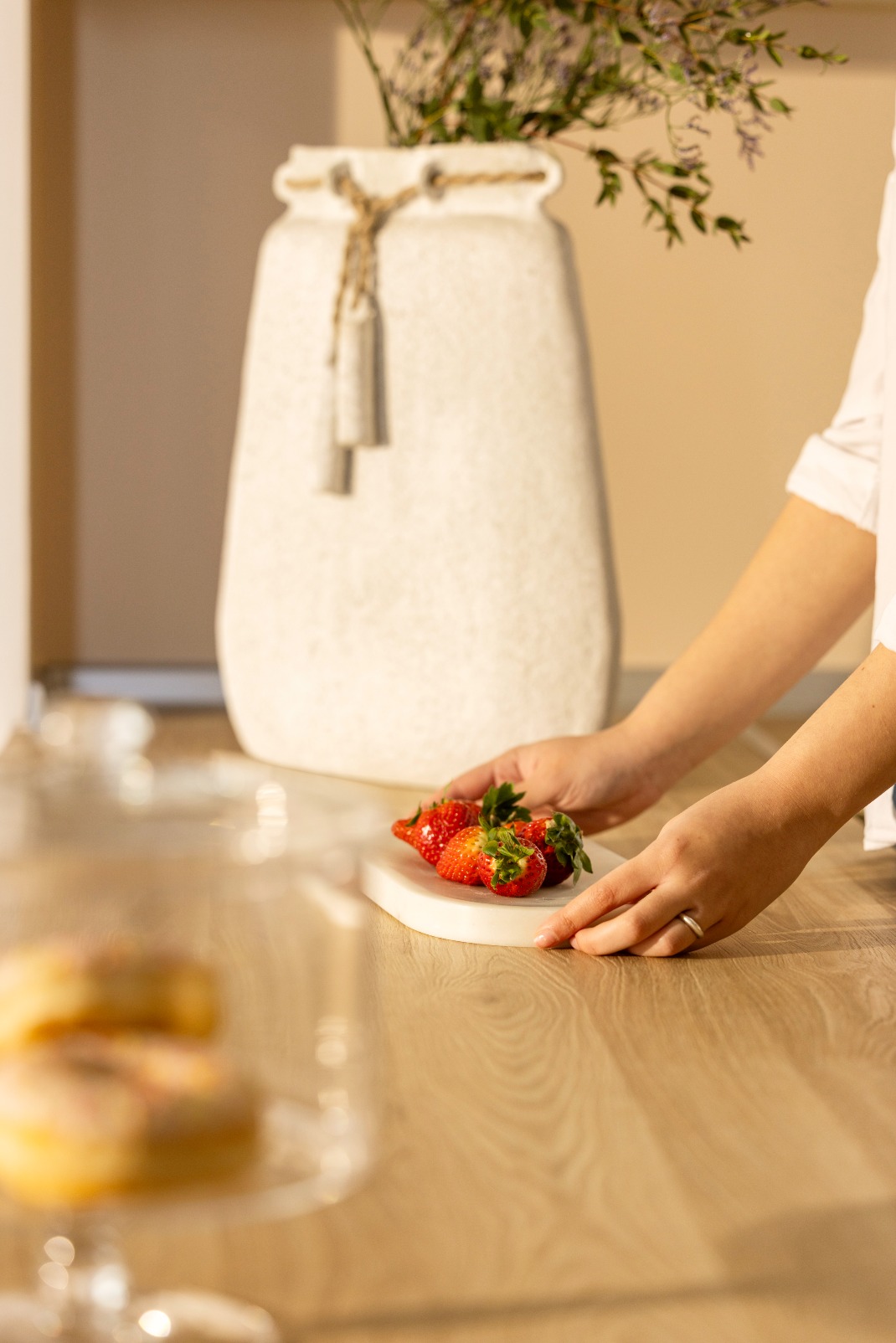 A person places a small white plate with fresh strawberries on a wooden table, with a large white vase and blurred pastries in the background.