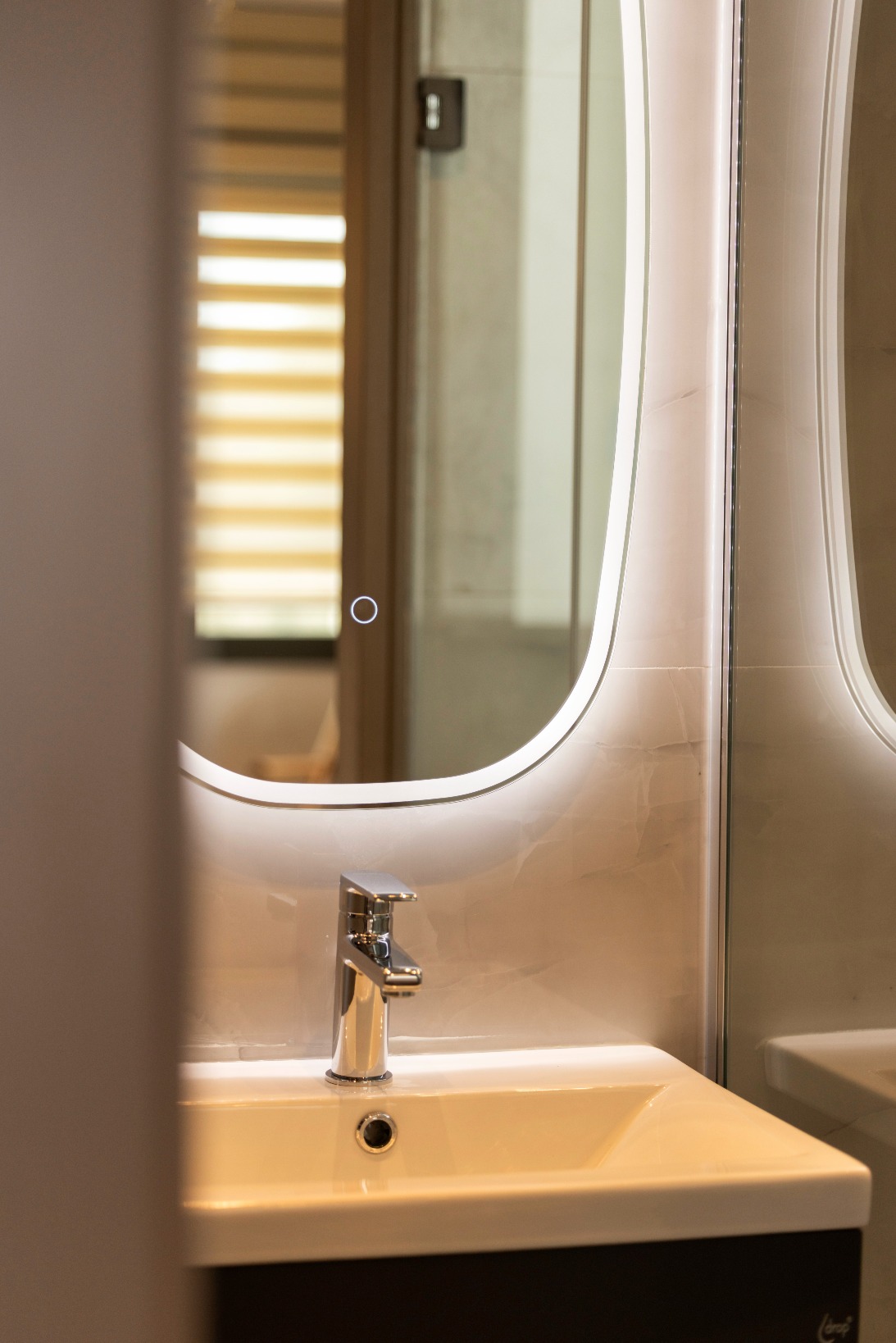 A modern bathroom basin with a chrome tap beneath a large, rectangular, backlit mirror. Blurred window blinds and part of a door frame are visible in the reflection.