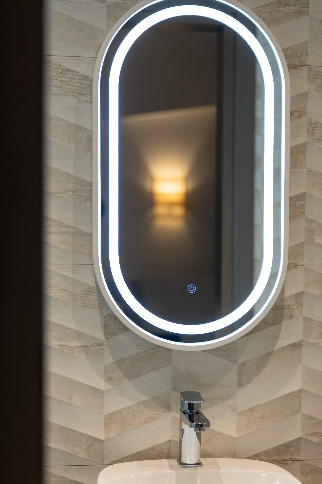 A modern bathroom with a geometric tiled wall, an oval-shaped LED-lit mirror above a white basin, and a chrome tap. The reflection in the mirror shows a blurred wall light fitting.