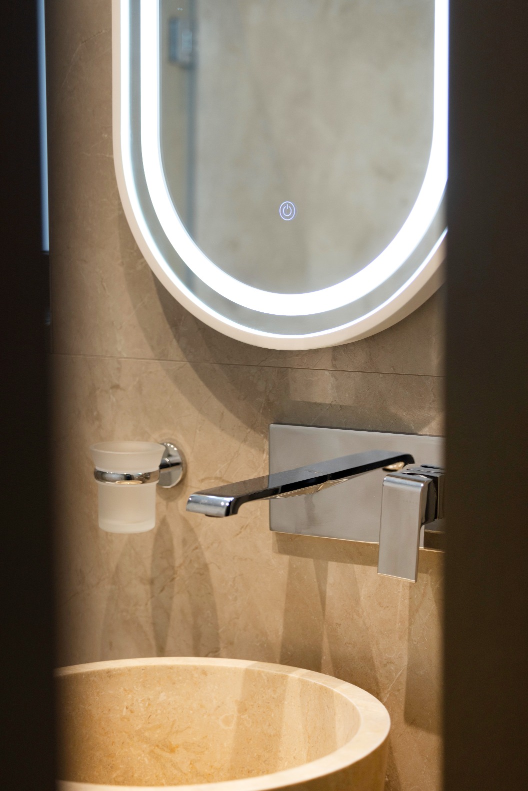 A modern bathroom basin with a round stone bowl, a wall-mounted tap, a glass tumbler holder, and an oval mirror with built-in LED lighting.