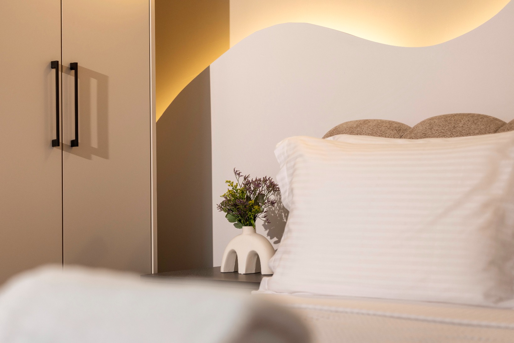 A close-up of a neatly made bed with white pillows, a bedside table holding a small decorative vase with flowers, and a wardrobe with black handles in a softly lit modern bedroom.