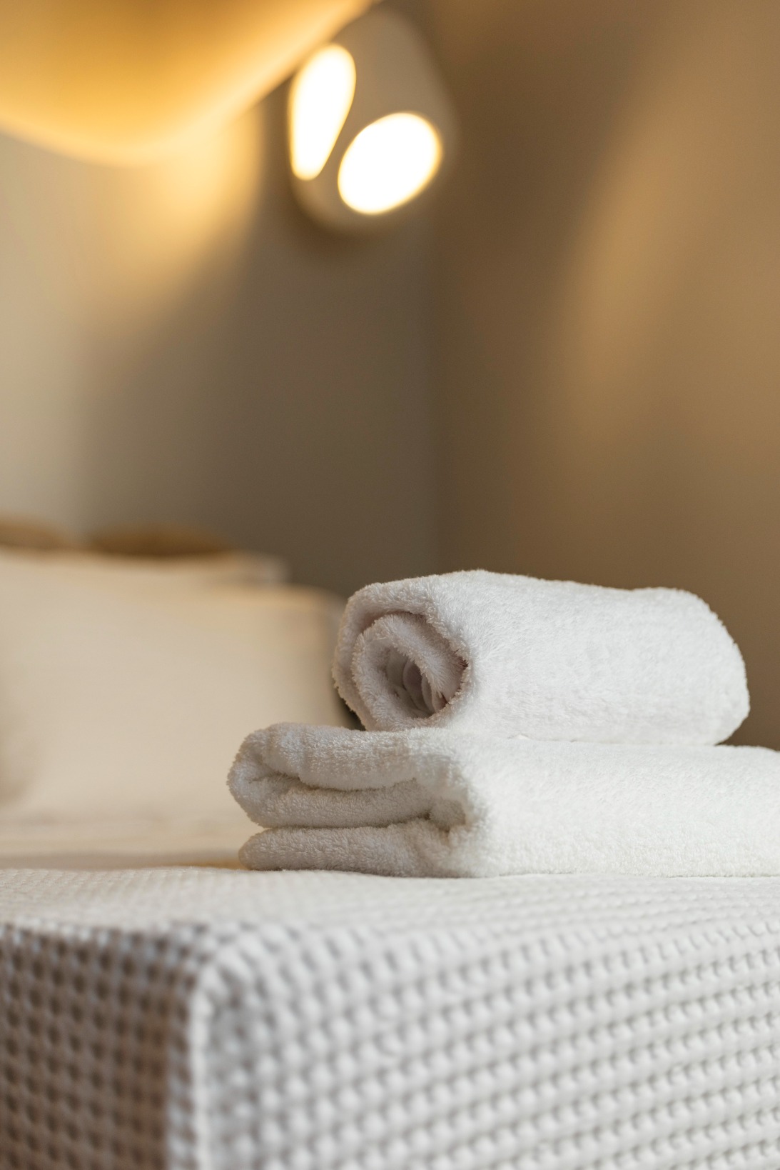 A close-up of two neatly folded white towels placed on a textured bedspread, with a softly lit bed and a modern wall lamp in the background, creating a cosy and inviting atmosphere.