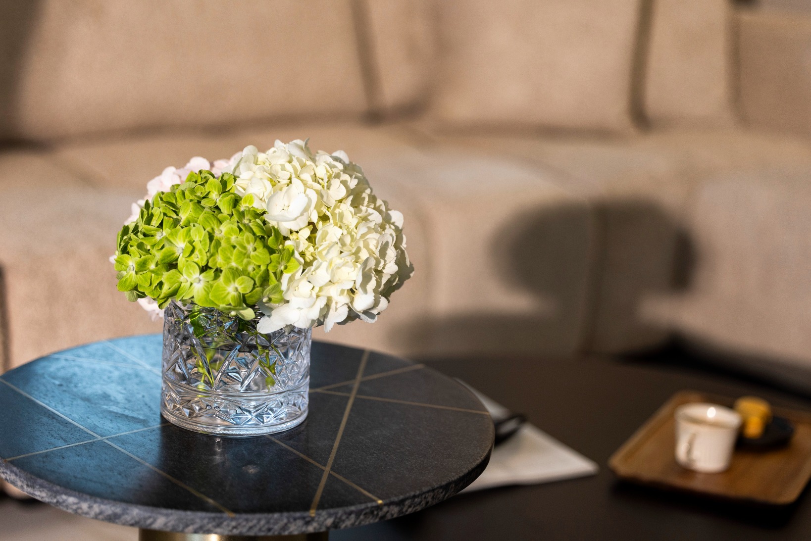 Crystal vase with white and green hydrangeas on a blue round table in a stylish living room.