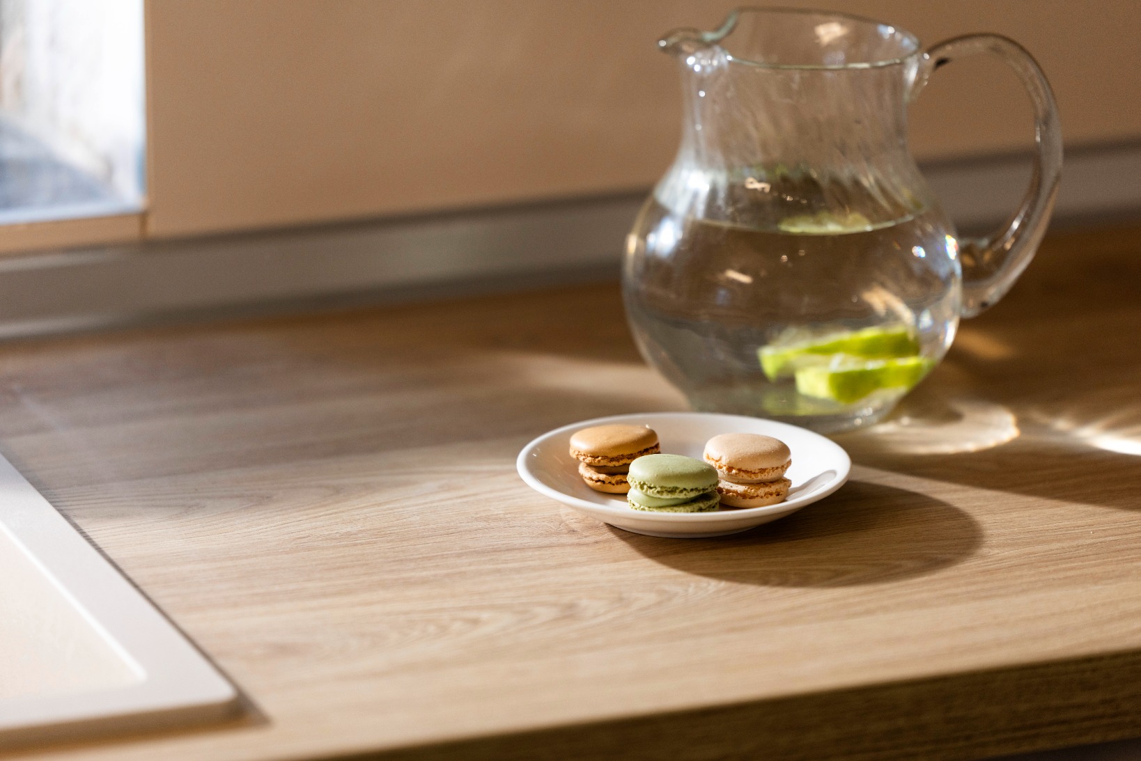 Plate of colorful macarons beside a glass water pitcher with lime slices on a wooden counter.