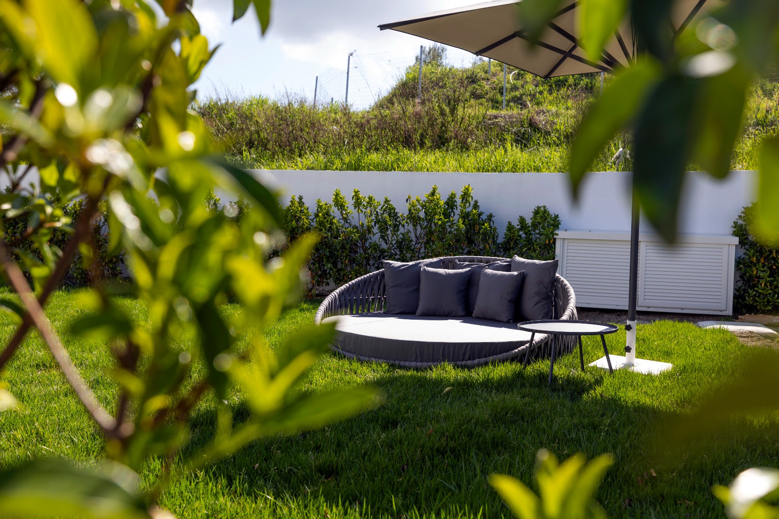 A cosy outdoor lounge area with a round daybed, grey cushions, a small side table, and a large umbrella, set on green grass with bushes and trees in the background.
