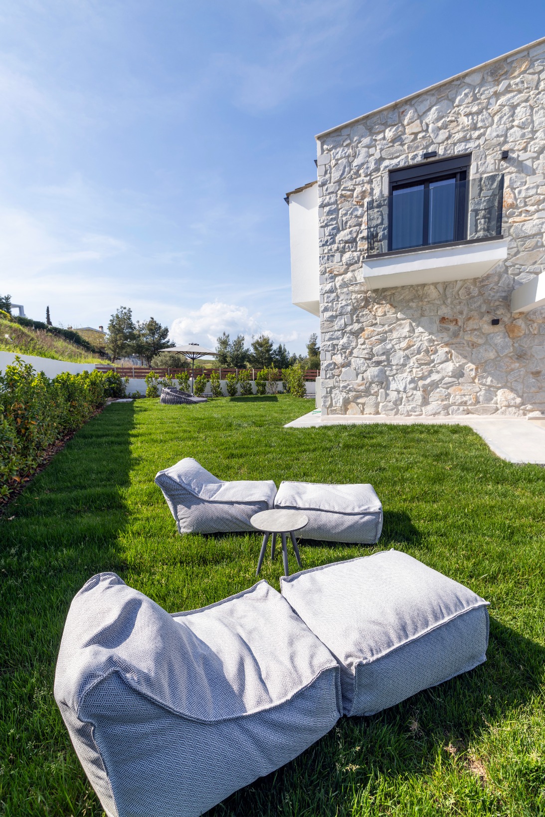 Two grey outdoor lounge chairs with a small round table between them are set on a green lawn beside a modern stone house under a blue sky. Shrubs line the edge of the garden.