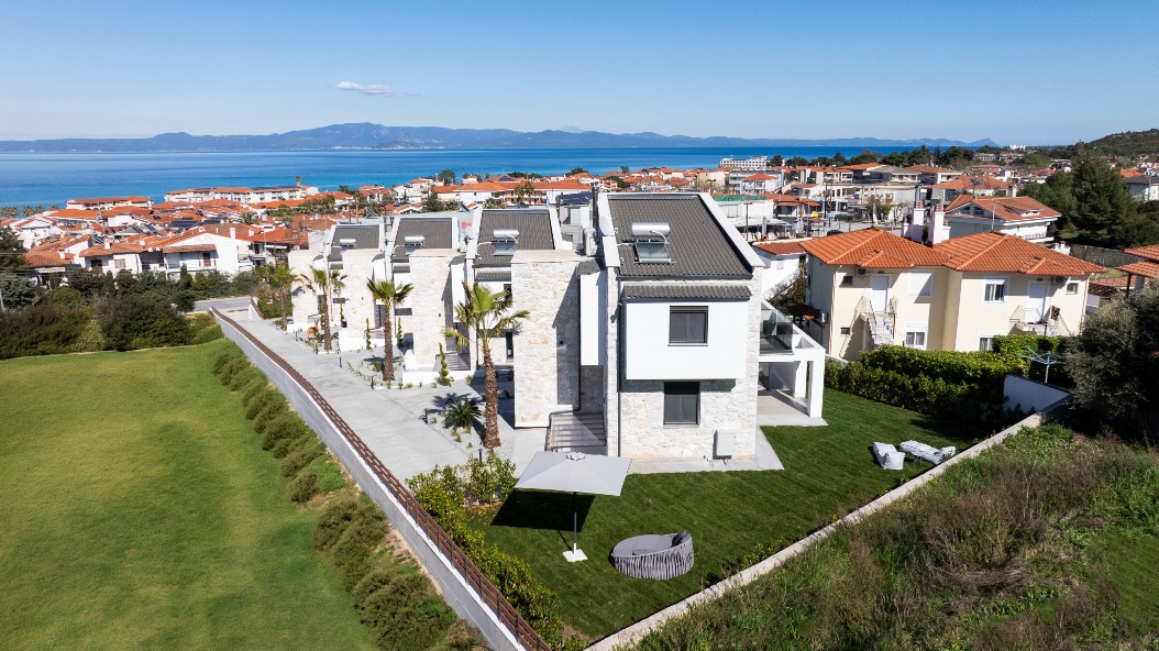 Aerial view of modern white villas with stone accents, set in a residential area near the coast. Red-roofed houses, blue sea, and distant mountains are visible under a clear sky.