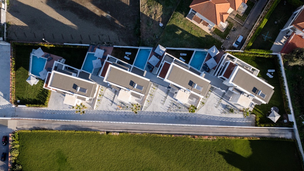Aerial view of five modern houses with swimming pools, aligned diagonally on a landscaped property, bordered by roads and green lawns, with neighbouring homes visible in the top right corner.