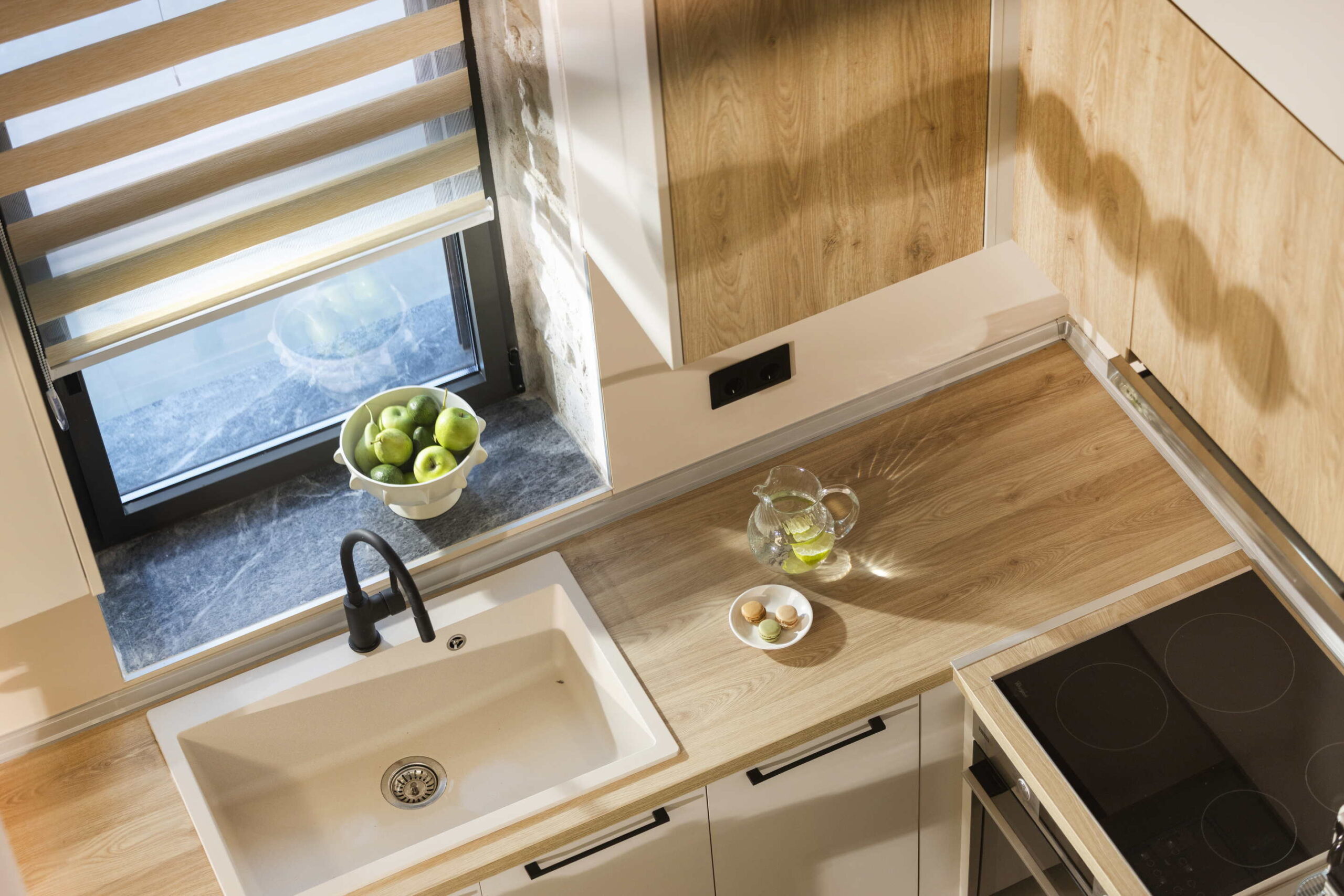 Overhead view of a modern kitchen with light wood cupboards, a white sink, black tap, a bowl of green apples on the windowsill, a glass jug of water with lemon, and a plate with three eggs on the worktop.