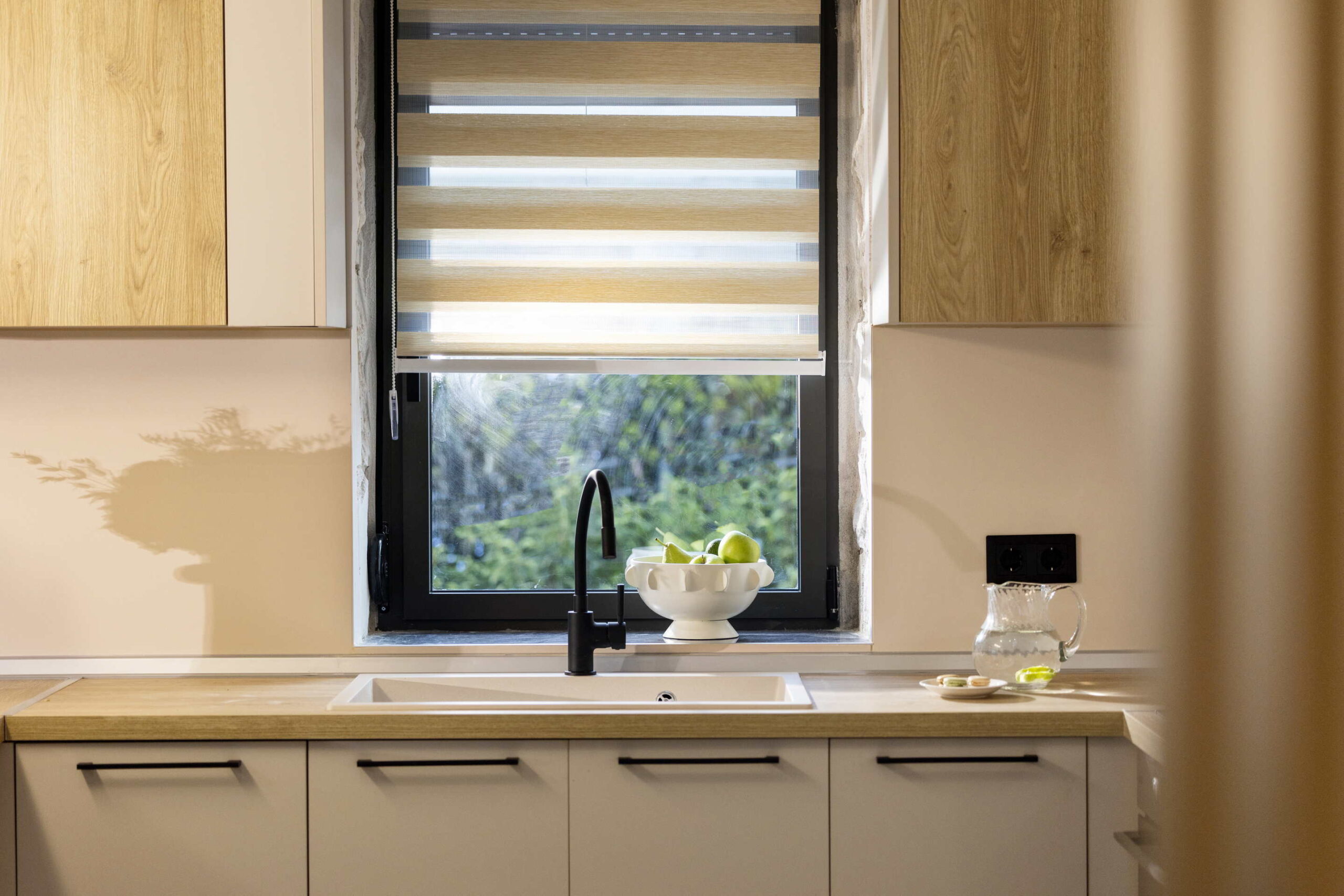 Modern kitchen with light wood cupboards and a white worktop, featuring a black tap, a bowl of green apples, a glass jug with water, and a plate with pastries near a window with striped blinds.