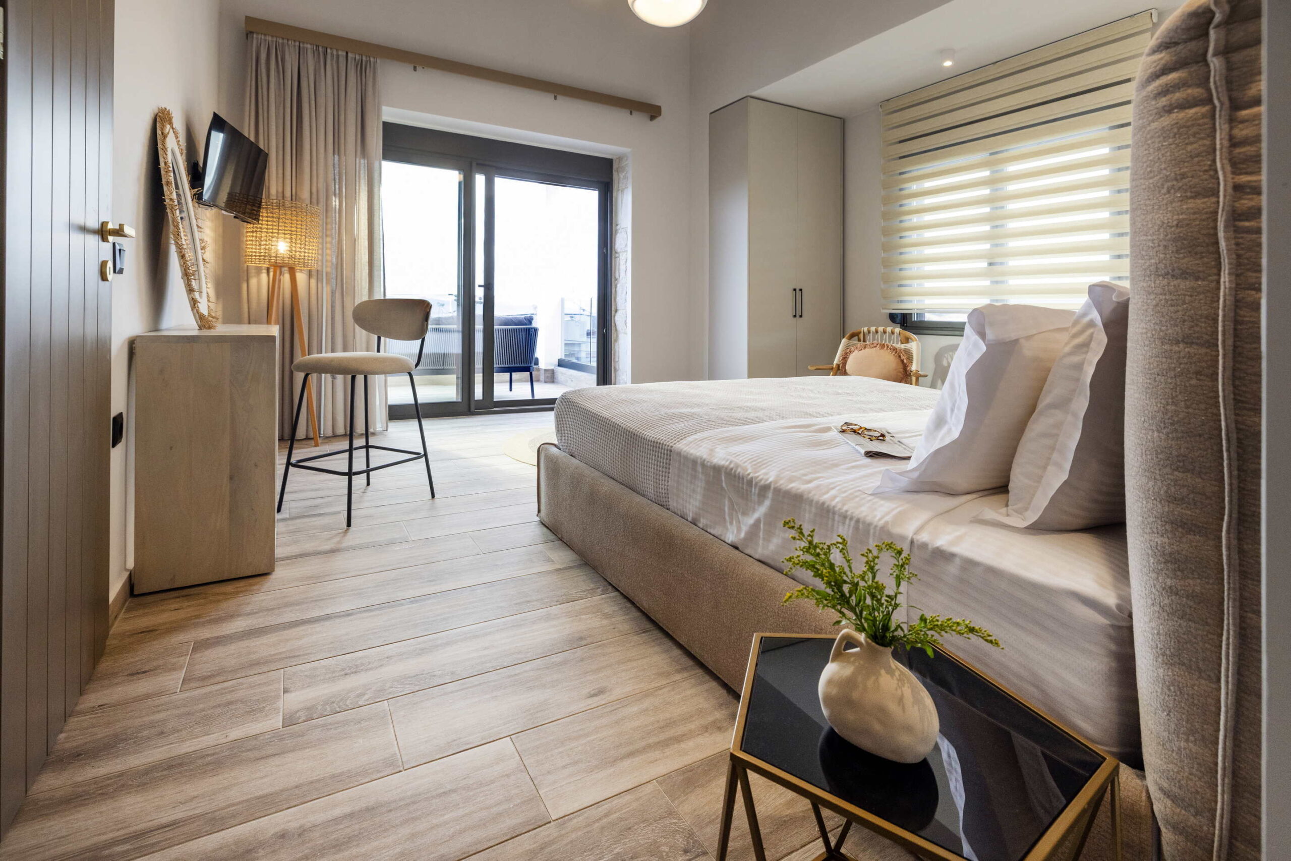A modern, cosy bedroom with a large bed, white linen, a small bedside table with a vase, a TV mounted on the wall, and floor-to-ceiling windows leading to a balcony with outdoor seating.