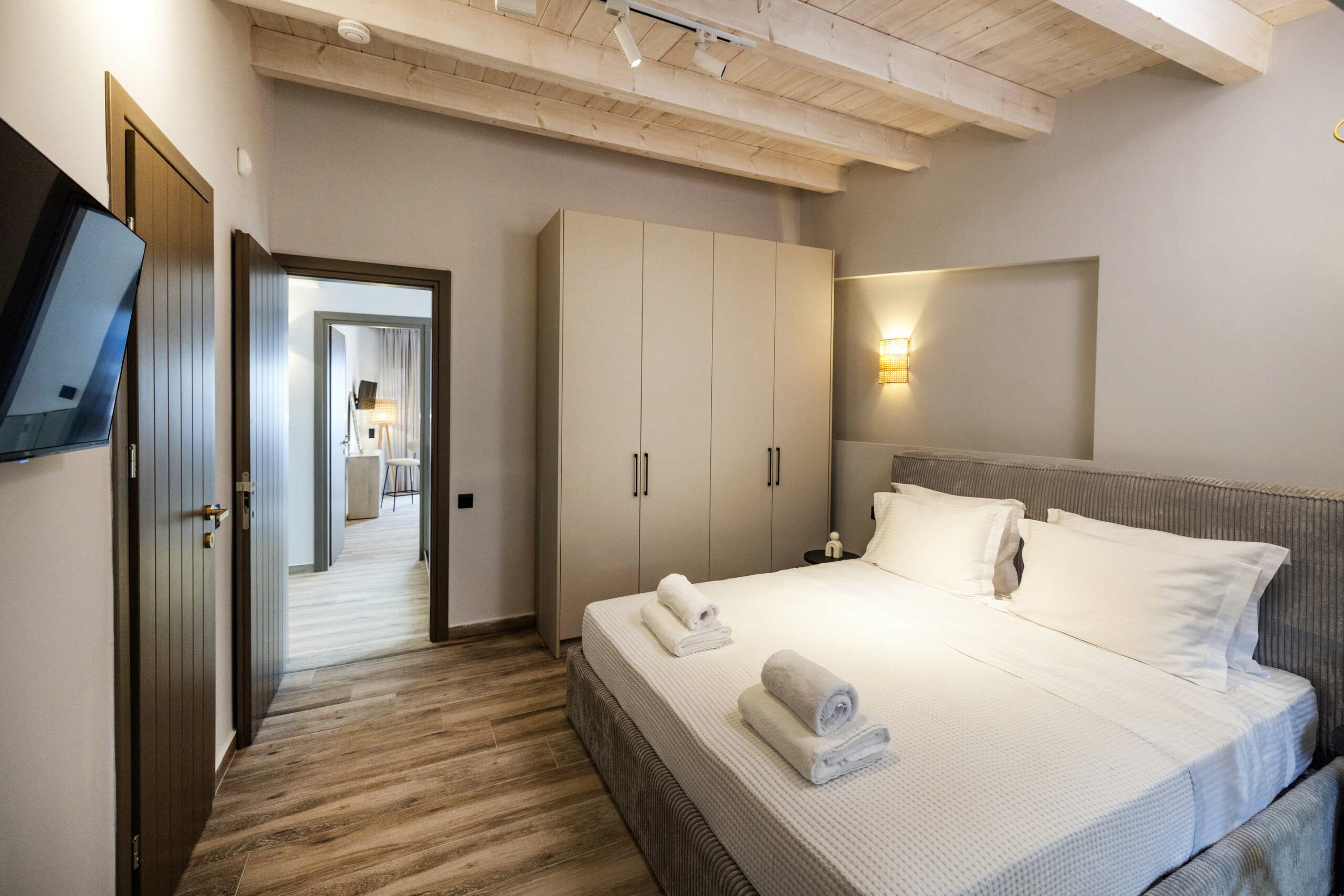 A modern bedroom with a neatly made double bed, white linen, towels on the bed, a large wardrobe, a wall-mounted TV, wooden floors, and an open door leading to a well-lit corridor.
