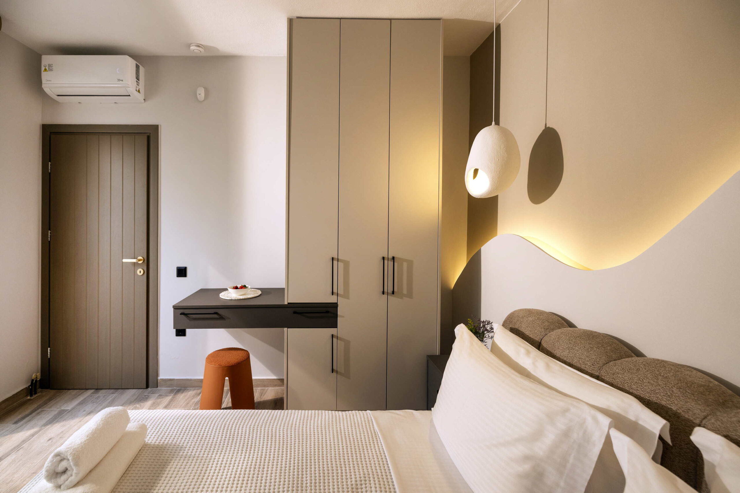 Modern hotel bedroom with neutral tones, large wardrobe, desk, orange stool, bed with white linens, and warm pendant light.