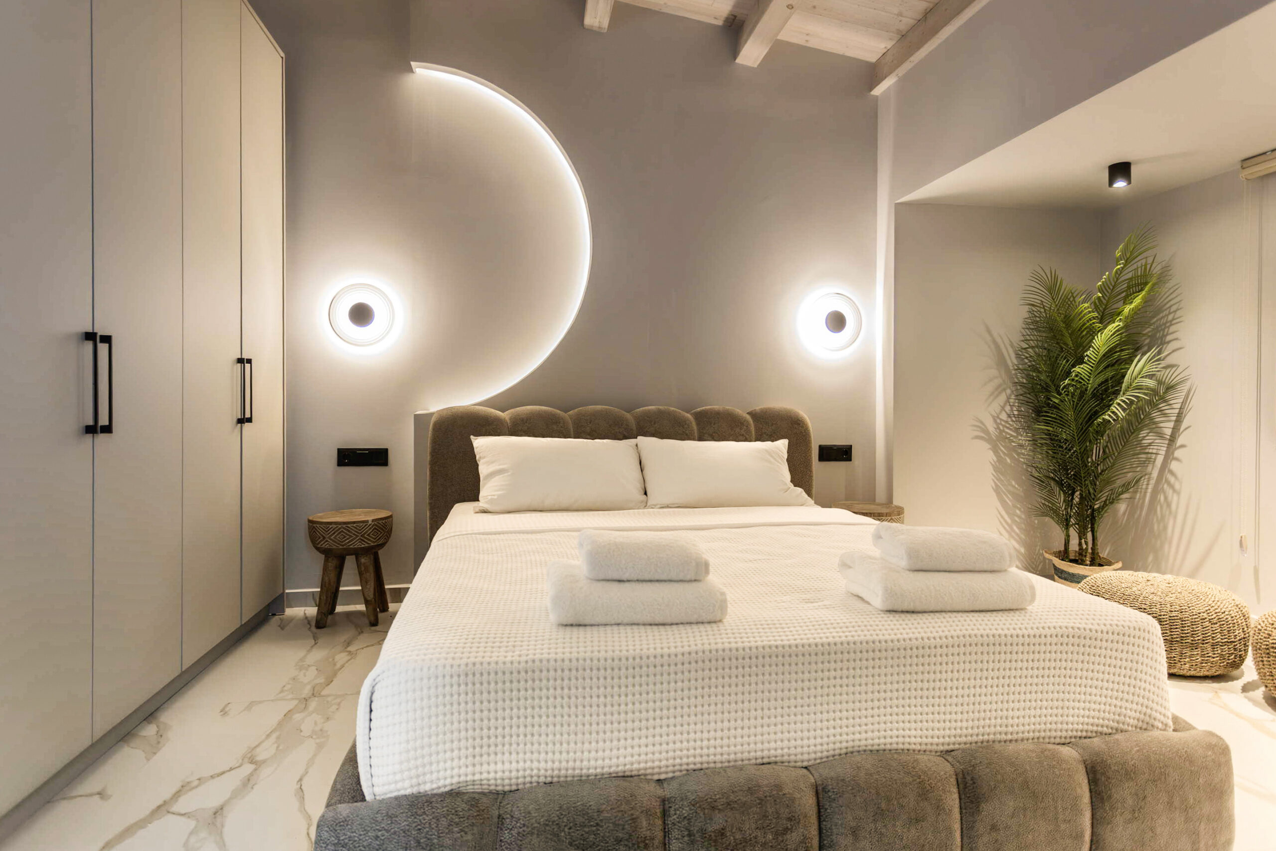Modern hotel bedroom with twin beds, ambient circular wall lights, neutral tones, and a potted plant.
