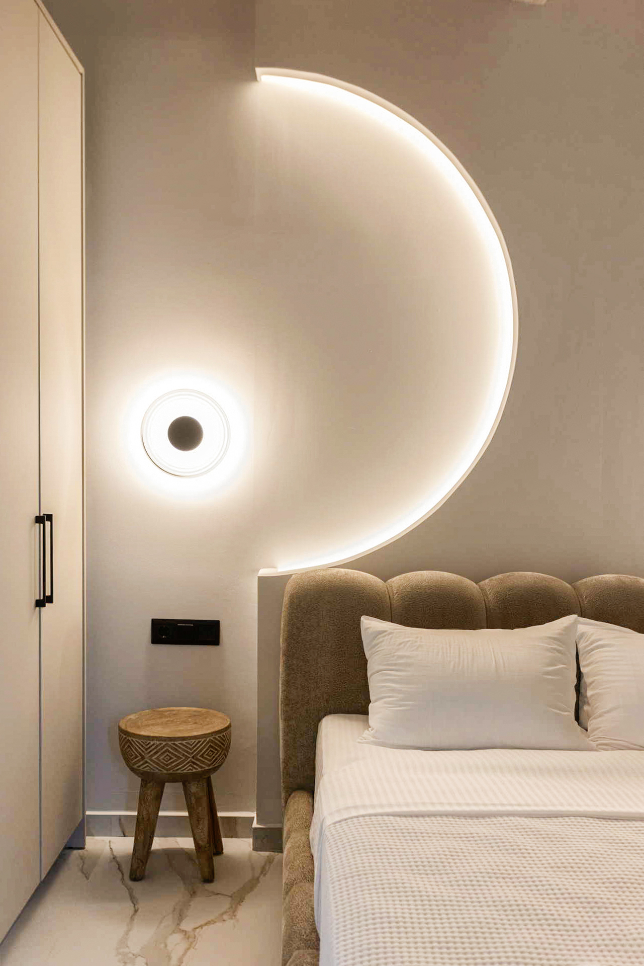 Modern hotel bedroom with circular wall light, beige upholstered headboard, white bedding, and marble floor.